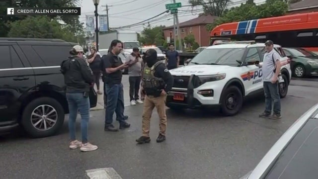 A masked ICE agent walks away from a police vehicle as spectators look on. 